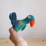 A vibrant blue parrot felt finger puppet is held by a hand against a bright background, in front of a simple wood surface. This handmade, needle-felted wool parrot finger puppet features a fluffy texture in shades of blue, green, and orange. Its distinctive orange beak and small eye emphasize its playful, handcrafted quality.