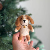 A handcrafted Cavalier King Charles Spaniel felt finger puppet is gently held in a hand, set against a warm, inviting backdrop with a Christmas tree. This unique wool felt puppet features characteristic brown, tan, and white colors, meticulously crafted with needle-felted details. It sports a black necklace with a golden bell, a sweet smile, fluffy ears, and an adorable black nose spot.