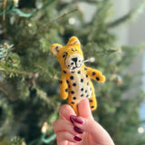 A handcrafted wool felt cheetah finger puppet, a delightful kids toy, is held by a female hand with red nail polish against a softly blurred evergreen tree with delicate lights. This charming cheetah puppet features a yellow body with distinctive black spots, a white muzzle, and tiny ears, showcasing its detailed fair-trade craftsmanship.