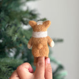 A charming handmade felt corgi finger puppet is held upright on a hand, positioned in front of a blurred Christmas tree, highlighting its festive appeal. This delightful felt toy for kids is showcased from the back, revealing its light brown fur, short limbs, and distinctive exaggerated puffy butt. The puppet features a white marking on its head, white paws, and a white band wrapped around its body.