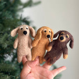 Three unique wool felt dachshund finger puppets, handcrafted in beige, khaki, and brown, are gently held in a female hand against a soft-focus backdrop of a Christmas tree and white wall. Each charming felted finger puppet features the breed's elongated body, floppy ears, and stitched facial details, bringing a playful and cozy mood to this display.