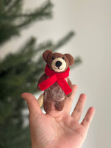 A handcrafted felt honey bear finger puppet is held upright in an open hand, with a blurred Christmas tree in the background. This unique, fair-trade honey bear puppet is crafted from brown and light gray felt, featuring a round head, small ears, and a light gray muzzle with a black nose. A bright red scarf is tied around its neck, adding a festive touch.