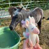 A handcrafted pastel unicorn felt finger puppet is held outdoors by a hand, positioned next to a real pony. This eco-friendly, needle felted unicorn puppet, made from 100% wool, features a pink snout, a winking eye with long lashes, and a soft rainbow mane in pastel shades.