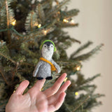 A handcrafted felt penguin finger puppet with headphones is held up by a female hand, set against a cheerful, blurred Christmas tree decorated with small lights. This unique felt finger puppet is gray with a white face and black feet. It wears green headphones and a thin yellow scarf, adding a festive touch.
