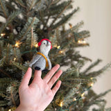 A unique, fair-trade felt penguin finger puppet with red headphones and a yellow scarf is held by a hand in front of a blurred, lit Christmas tree. This charming gray and white handmade toy, crafted in Nepal, brings a festive, playful touch for kids.