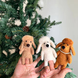 Three handcrafted felt poodle dog finger puppets are presented by a female hand against a festive Christmas tree adorned with pine cones. Each unique felt poodle dog puppet, in white, beige, and brown, features fluffy hair and a delicate plastic pearl necklace, perfect for imaginative finger play.