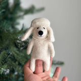 A white poodle dog felt finger puppet, held in a female hand, is displayed against a blurred festive Christmas pine tree background. This charming wool felt poodle finger puppet features long, floppy ears and a delicate plastic pearl necklace. Its fluffy texture and simple design evoke a handmade charm and playful sophistication, ideal for kids' finger puppet play.