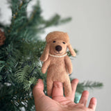 A beige handmade felt poodle dog finger puppet is held by a female hand, presented clearly against a softly blurred Christmas tree backdrop. This unique finger puppet features a delicate plastic pearl necklace and fluffy hair atop its head, highlighting its charming, handcrafted details.