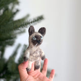 A handcrafted felt Siamese cat finger puppet is held by a female hand against a bright, cheerful backdrop with a blurred Christmas tree. The plush felt puppet features a light tan body with dark brown markings on its face, ears, and paws, blue eyes, and white whiskers. This unique, fair-trade felt toy for kids is ethically made, adding charm to playtime.