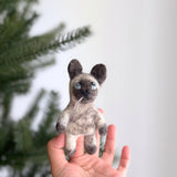 A handmade Siamese cat felt finger puppet is held in a female hand, with a blurred Christmas tree in the background, creating a festive mood. This unique, needle felted puppet showcases the distinctive pointed coloration of a Siamese cat, featuring darker ears, paws, and face contrasting its lighter body. It has charming blue eyes and delicate white whiskers. Crafted from plush felt by a Fair Trade artisan collective in Nepal, this eco-friendly toy supports imaginative play.