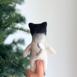 A female hand presents a meticulously crafted felt tuxedo cat finger puppet against a bright background with a Christmas tree visible to the left. This plush felt tuxedo cat finger puppet, seen from behind, features a black head and a white body with a tail and outstretched arms, showcasing its textured appearance. This unique, handmade felt toy is ethically produced by a Fair Trade artisan collective.