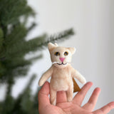 A female hand holds a handcrafted White Siamese Cat felt finger puppet, displayed against a soft white background with a Christmas tree edge visible. This charming felt finger puppet features small upright ears, a pink nose, a black stitched mouth, white whiskers, and yellow eyes. Its slender white body includes short arms and legs, with a tan tag at the back. This unique felt toy is a fair-trade piece, reflecting ethical craftsmanship.