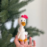A handmade white felt duck finger puppet, part of a honeymoon couple set, is held by a hand against a blurred Christmas tree background. This adorable felt finger puppet features an orange beak and feet, a pink bow on its head, and a strand of white beads around its neck, showcasing its unique, handcrafted design.