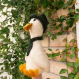A handcrafted felt duck finger puppet is playfully held on a female hand, set against lush green foliage climbing a brick wall. This whimsical, off-white needle felted duck features a black cap and scarf, bright orange feet and beak, and striking blue eyes. Its intricate detailing highlights the quality craftsmanship, making it ideal for imaginative play.