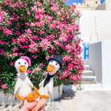 Two handcrafted felt duck finger puppets, portraying a whimsical duck couple, are held by a female hand against a vibrant backdrop of pink flowers and white buildings. The white felt duck on the left wears a pink hat and white collar, while the one on the right sports a black hat and scarf. Both adorable duck puppets feature orange beaks and feet, highlighting their charming, expertly crafted details.