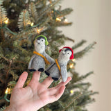 Two playful felt penguin finger puppets, part of a unique set of two, are held by a female hand against a festive Christmas tree background. These handmade felt puppets each have a gray body, white face, and yellow scarf. One penguin wears green headphones, while the other wears red headphones, highlighting their charming and detailed craftsmanship.