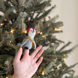 A handcrafted wool felt penguin finger puppet sits on a female finger, displayed against a blurred Christmas tree adorned with warm lights. This grey and white felt penguin finger puppet features playful red headphones and a yellow scarf, highlighting its unique design and supporting female artisans in Nepal.