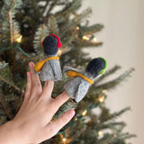A female hand holds a set of two charming, handcrafted felt penguin finger puppets against the soft glow of a Christmas pine tree. Each gray and black felt penguin features a distinctive yellow band around its body, with one wearing red headphones and the other green. These unique, playful finger puppets are perfect creative toys for kids.
