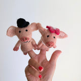 A pair of handmade pink piggy felt finger puppets are playfully displayed on a female hand with red nail polish against a bright white background. These unique needle-felted piggy puppets feature textured wool bodies, perky ears, tiny black eyes, and smiling mouths. One piggy puppet wears a black top hat, while the other is adorned with a pink flower and pearl beads around its neck, highlighting their whimsical, handcrafted charm.