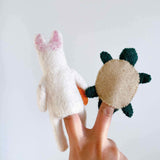 A set of two handmade felt finger puppets, a white rabbit and a tan turtle from The Tale of the Turtle and the Rabbit, is presented by a hand against a plain white background. The white felt rabbit finger puppet features pink-spotted ears and an orange carrot detail. The tan felt turtle finger puppet displays dark green legs. These handcrafted felt puppets are perfect for imaginative storytelling.