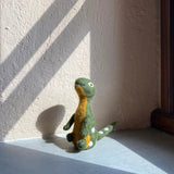 A handcrafted Brontosaurus felt finger puppet, from the 'Dino Brothers' collection, rests on a light gray surface near a wall, naturally lit by sunlight creating subtle shadows. This unique needle-felted dinosaur puppet, made from 100% wool, features a long neck, a green body with an orange underbelly, and small white dots along its back and tail. Its detailed stitching and small eye highlight its handmade, 3D design for a lifelike appearance.