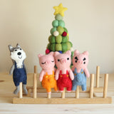 Four handmade felt finger puppets, depicting the Three Little Pigs and a Big Wolf, are displayed on a wooden stand. A decorative felted Christmas tree stands behind the puppets against a plain wall. The wolf puppet is on the left, facing the three pig finger puppets, each wearing unique colored overalls and showcasing distinct facial details for imaginative play.