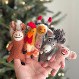 A female hand presents a festive set of five handcrafted wool felt farm animal finger puppets against a blurred Christmas tree background. This charming collection features a brown wool felt horse with a white mane, a white and orange hen, a white and black cow with a small bell, a grey sheep, and a pink pig. Each puppet features stitched smiles, highlighting their unique craftsmanship.