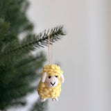A yellow mini felt fluffy sheep ornament hangs from a branch, presented against a soft, neutral backdrop with a hint of festive greenery. This handmade felted wool ornament features a fluffy texture, small white ears and feet, and a minimalist stitched face. It is part of a set of unique pastel-toned felt sheep ornaments, bringing cozy, handcrafted holiday warmth.
