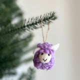A purple felt fluffy sheep ornament hangs from a green Christmas tree branch. This mini felted wool sheep ornament features a soft, fluffy texture, small white ears, and is suspended by a jute string, adding a playful touch to holiday decor.