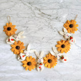 A cheerful felt Bunny and Sunflower garland is displayed in a semi-circular shape on a white marble surface. This 36-inch long felt garland features 6 alternating bright yellow sunflowers with dark brown centers and 6 white bunnies. Each white bunny playfully holds a small orange carrot, adding a delightful touch.