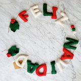 A festive "Feliz Navidad" wool felt garland is arranged in a semi-circular shape on a white marble surface. It features plump letters in red, white, and green spelling "Feliz Navidad," connected by a jute string. Decorative green holly leaves with small red berries adorn both ends, with a small product tag visible near one letter.