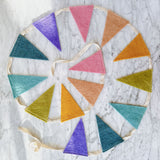 A vibrant felt flag garland, a festive party banner, is arranged in a circular pattern on a white marble surface. This handmade felt garland features numerous triangular flags in rich shades of purple, pink, orange, yellow, green, and teal, all connected by a white string, showcasing its simple yet cheerful design.