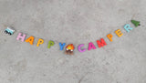 A Happy Camper felt garland is displayed on a textured gray surface. It features a blue minivan, a felt campfire, and a green tree. The garland spells "HAPPY CAMPER" with individual felt letters in pink, orange, yellow, green, blue, purple, and red, strung together on a thin brown twine.