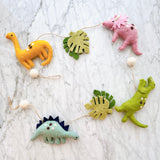 A vertical Jurassic Dinosaur felt garland is playfully displayed on a marbled surface. This wool felt garland features a yellow brontosaurus, a pink triceratops, a green T-Rex, and a light blue stegosaurus, each with small felt spots. The dinosaurs are connected by a thin string interspersed with wool balls and separated by green monstera leaves, with a charming copper bell at one end.