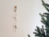 The felt seal garland hangs against a stark white wall in a calm, minimalist setting, with a Christmas tree gently visible nearby. This charming felt garland features three seals, alternating in light gray and white colors.