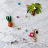 A playful felt garland, featuring a tropical trio of a pink flamingo, a yellow pineapple, and a palm tree, is arranged against a white marble backdrop. The pink felt flamingo has a white heart and black beak, with a small copper bell at its end. A yellow felt pineapple with green leaves is in the center, and a felt palm tree with a brown trunk, green leaves, and white coconuts completes the trio. All elements are connected by a string of colorful felt balls, creating a cheerful mood.