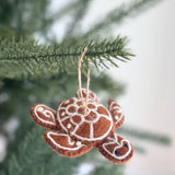 A brown wool felt turtle ornament hangs by a jute string from a blurred green Christmas tree branch. This felt gingerbread turtle ornament features white decorative stitching on its shell and flippers, resembling a charming gingerbread cookie. Tiny black bead eyes complete its whimsical look.