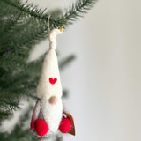 A handcrafted needle felted gnome with love ornament hangs on a Christmas tree branch. The gnome features a creamy white, cone-shaped wool felt hat with a small red heart and a tiny gold hanging bell. Its body is made of textured light and dark gray wool felt with small red feet, and a round tan nose peeks from under the hat, adding whimsical charm.