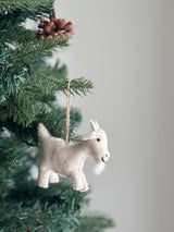 A soft, off-white felt goat ornament, suspended by a jute string, hangs on a vibrant green Christmas tree. The felt goat ornament features a fluffy beard and perky ears, lending a rustic and warm feel to the festive tree decorated with needles and a pine cone.