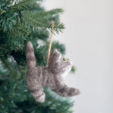 A gray American Short Hair cat felt ornament hangs on a Christmas tree. This cozy wool felt ornament is hand-stitched with intricate embroidery detailing its whiskers, ears, tail, and limbs. A jute string allows for easy hanging.