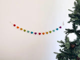 A cheerful felt garland, featuring seven rainbow-hued pastel stars strung on a cotton thread, is displayed against a white wall, secured by a clear hook. An artificial pine tree stands to its side, enhancing the festive atmosphere.