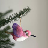 A needle felted wool felt hummingbird Christmas ornament is suspended from a pine branch against a light background. This unique Christmas ornament features a white belly, a dark beak, and a jute string loop at the top, ready for display.