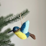 A vibrant needle felted hummingbird Christmas ornament, crafted from 100% wool felt, is delicately suspended from a pine branch. This three-dimensional hummingbird ornament showcases a textured design in shades of blue, yellow, and brown, ready for hanging.