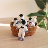 Wool felt astronaut keychains are artistically arranged inside and beside a woven brown basket, resting on a light wooden surface. These handcrafted keychains are primarily white, featuring black helmets, gray gloves and boots, and small dark square accents. One unique keychain also displays a tiny American flag, with each having a metal key ring attached.
