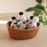 Multiple handcrafted wool felt astronaut keychains are nestled in a woven basket on a light wooden surface, with a hint of a green plant in the background. Each charming felt astronaut features a round white body, a black helmet, and gray arms. Some of these unique keychains include a small American flag design on their bodies, each attached to a metal keychain.