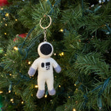 A handcrafted wool felt astronaut keychain hangs playfully on a lush, green Christmas tree, which is adorned with warm white lights. The stylized white astronaut features a black visor, gray hands and feet, and small black squares on its body. It is attached to a metal keychain with key ring.