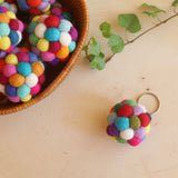 Colorful wool felt ball pompom keychains are displayed on a light wooden surface. A woven basket holds a cluster of these handcrafted keychains, while one rests nearby, attached to a metal clasp. Made from many smaller felt balls, they feature vibrant hues of red, blue, yellow, pink, green, and white, creating a playful effect.