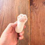 A female hand gently holds a handmade felt cat paw keychain against a rustic wooden backdrop. This unique keychain is a 1:1 replica of a cat's paw, featuring a white base with puffy, textured pink paw pads. A metal clasp is attached, emphasizing its tactile appeal and handcrafted detail.