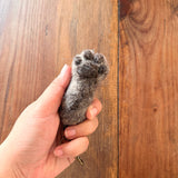 A gray wool felt cat paw keychain is gently held by a female hand against a rustic wood background. This handmade felt keychain replicates a cat's paw with detailed puffy toes. A copper keychain with a clasp dangles from the base of the paw, conveying a warm, comforting feel.