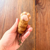 A female hand gently holds a textured orange and brown wool felt cat paw keychain against a warm wood surface. This unique wool felt keychain is shaped like a cat's paw with puffy, pink toe beans, and includes a copper keychain with a clasp. The playful design evokes a sense of comfort.